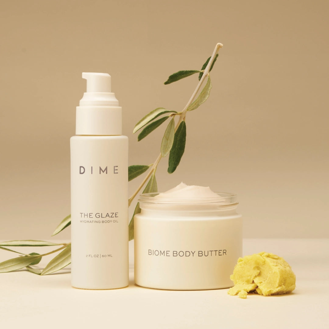 Two white DIME skincare products: hydrating body oil pump bottle and body butter jar, displayed with green botanical stems an