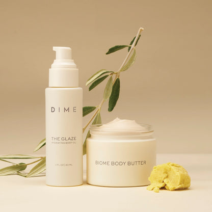 Two white DIME skincare products: hydrating body oil pump bottle and body butter jar, displayed with green botanical stems an