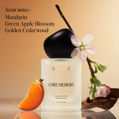 White cylindrical perfume bottle with black cap, mandarin peel, and white blossoms on branch against tan background.