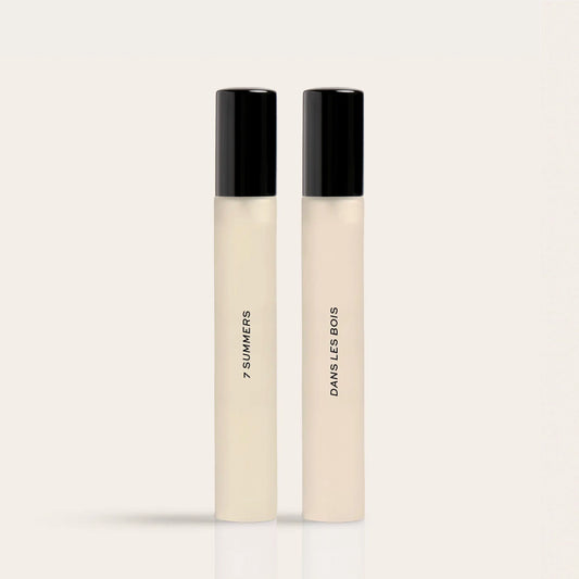 Two cylindrical fragrance bottles with black caps and cream-colored liquid, labeled "7 Summers" and "Damselless Bodies".