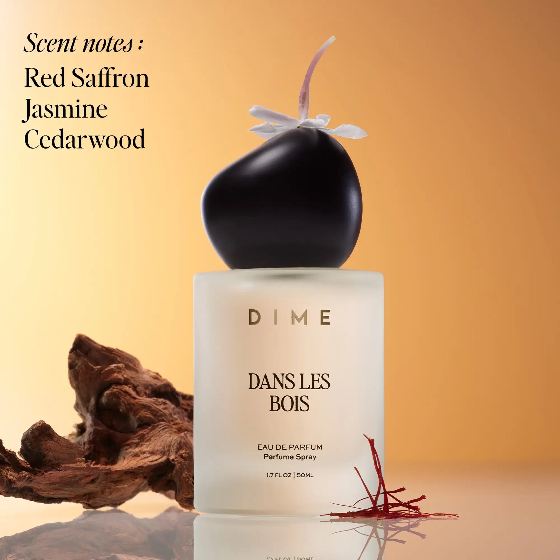 Black and white perfume bottle labeled "Dans Les Bois" with purple ribbon bow, surrounded by wood and saffron threads on tan 