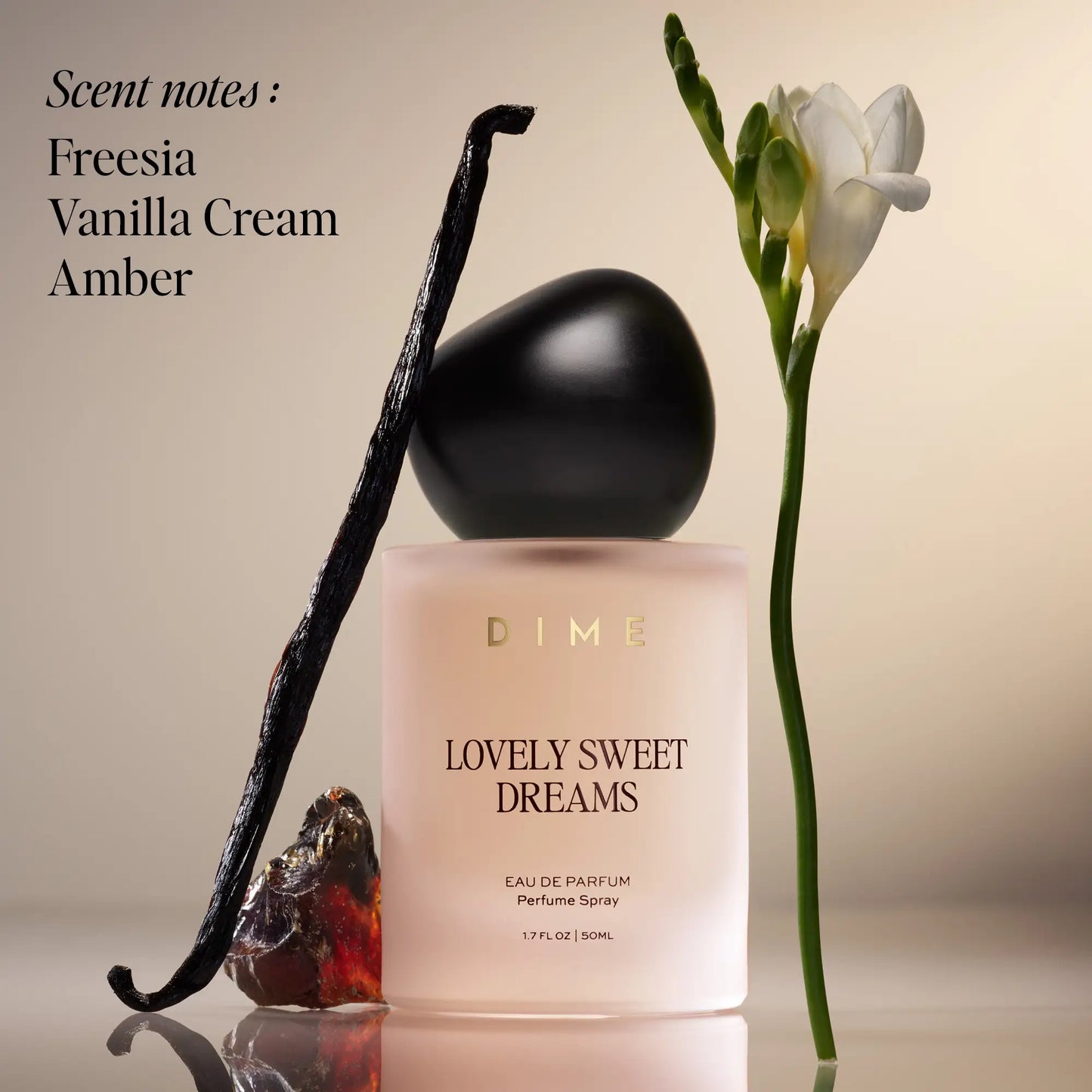 Pale pink perfume bottle with black spherical cap, vanilla bean pod, and white freesia flower on neutral background.